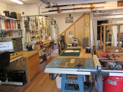 View from the garage door looking in to the main part of the shop. Island to the right, and benches along the left wall. You can see the tables saw's blast gate is open and the duct going up (power coming down)