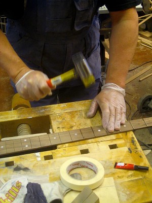 After installing side markers I cut the frets and hammered them into their slots. I mask the slot edges and put a few drops of CA glue into the slot prior to installing the fret. I have also made a radiused fret pressing caul to use with the drill press, but I'm more comfortable doing it this way.